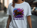 White t-shirt with brown dog running through blue and pink paint splatters, text "Unleash the Joy".