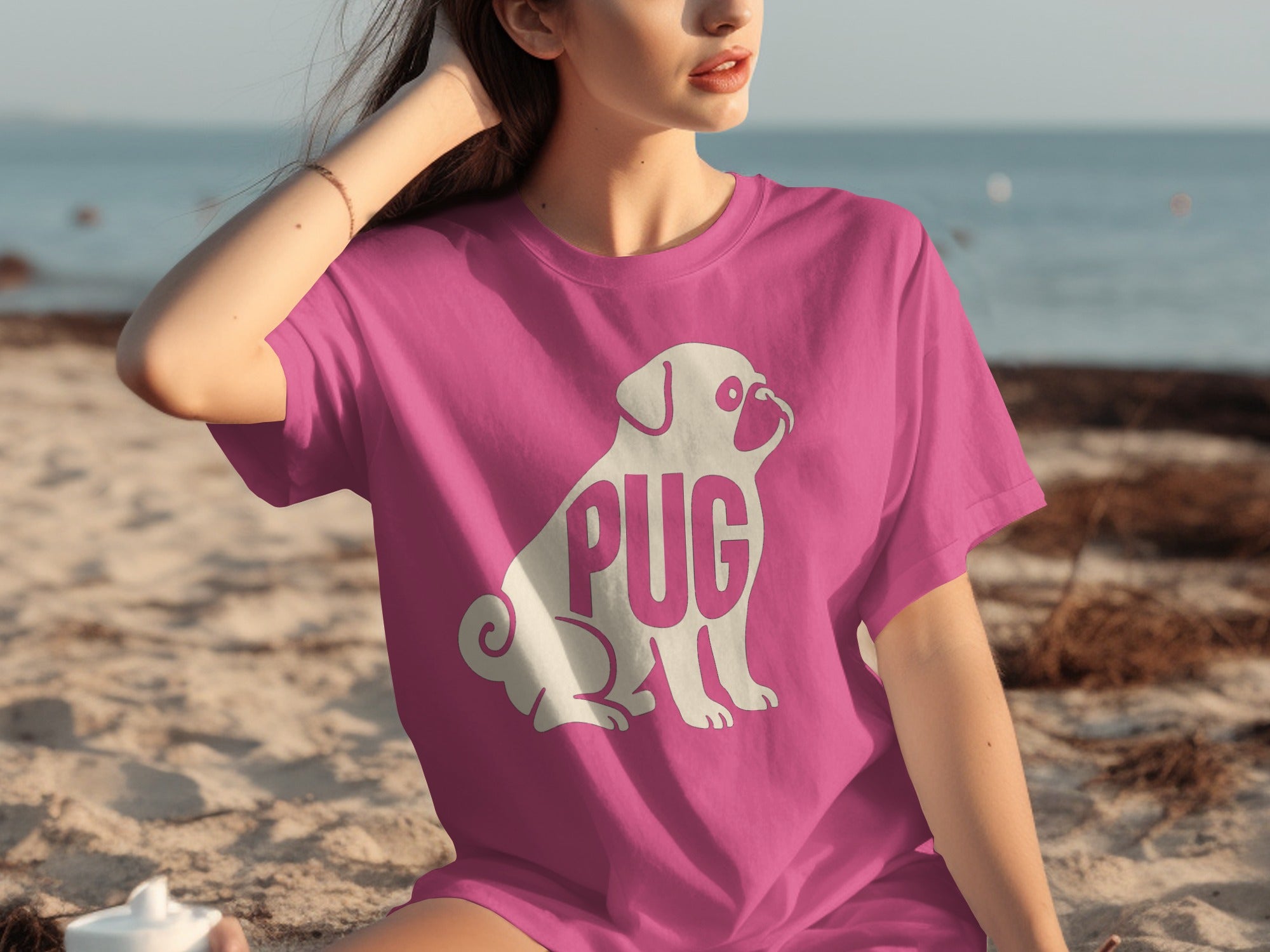 Pink t-shirt with a white pug graphic and "PUG" text on a beach.