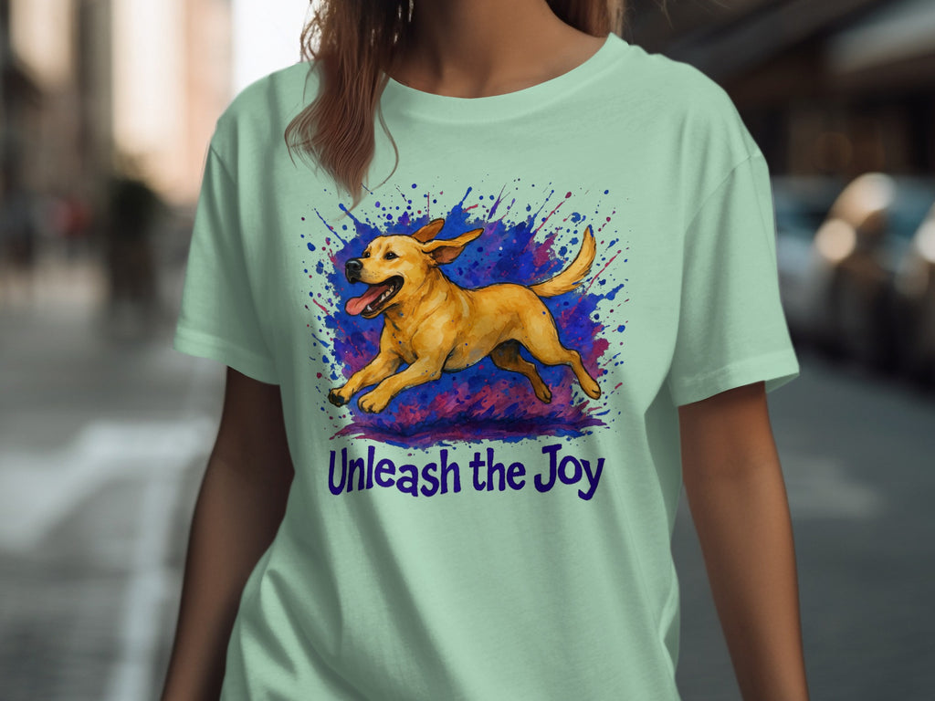 Light green t-shirt with watercolor print of a yellow dog running through colorful splatters and text "Unleash the Joy".