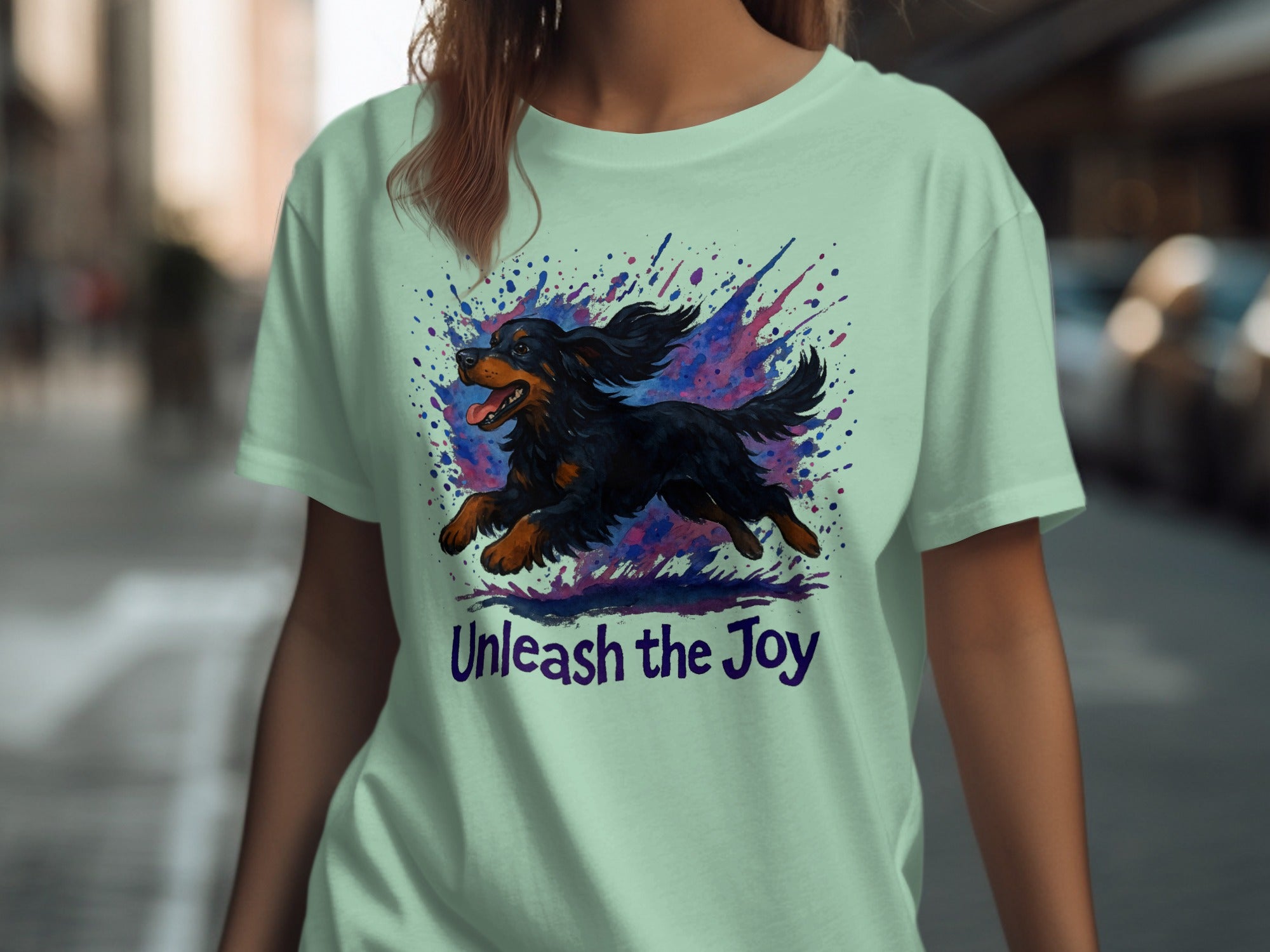 Light green t-shirt with graphic of a black and tan dog running amid colorful splatters and text "Unleash the Joy".