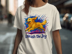 White t-shirt with a golden retriever running through colorful paint splatters, text "Unleash the Joy".