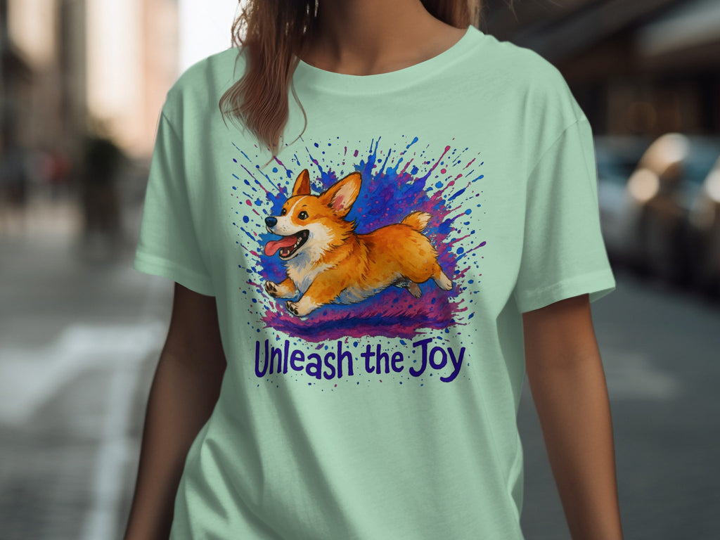Light green t-shirt with a corgi jumping through colorful paint splatters and "Unleash the Joy" text.
